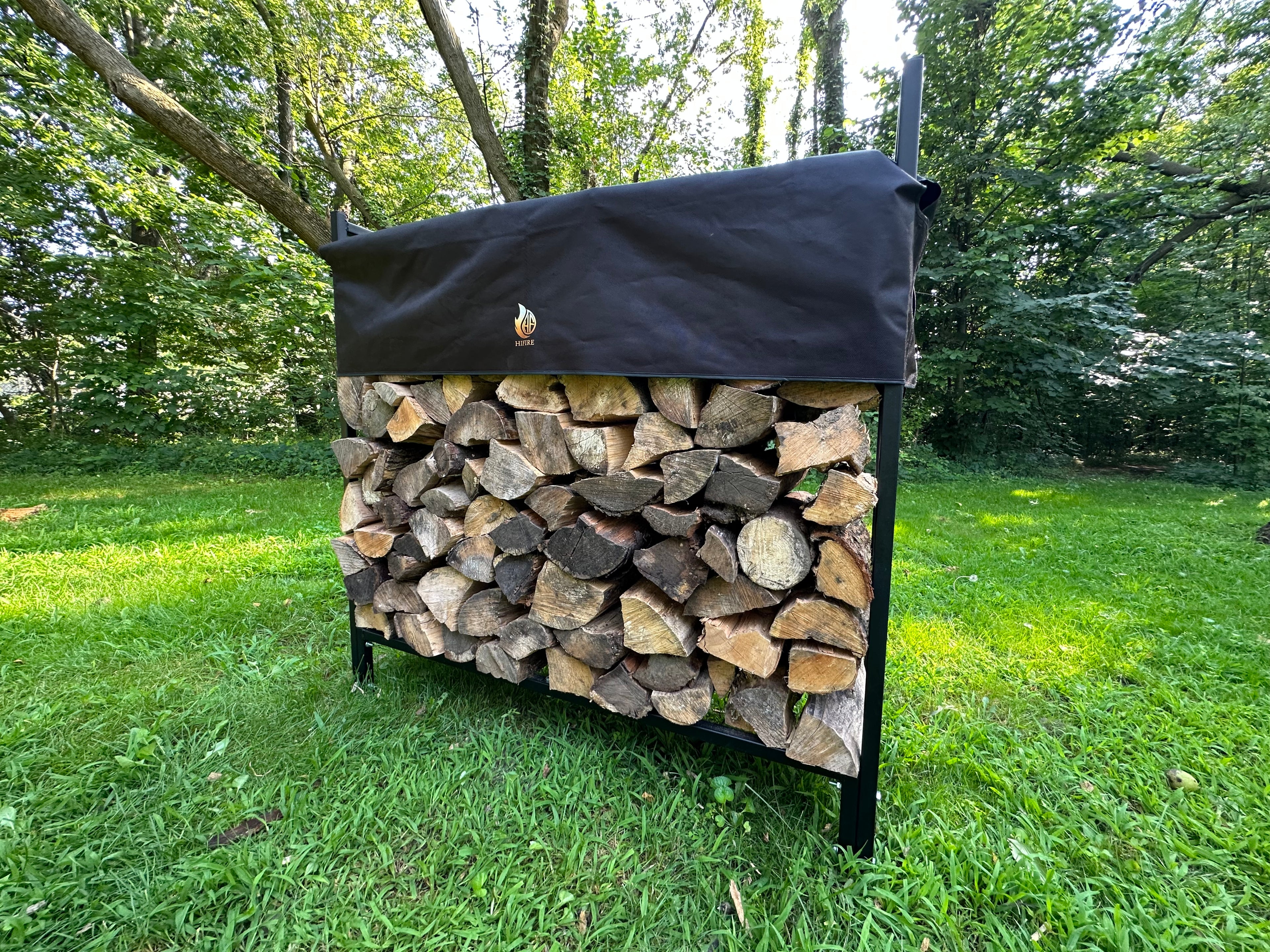 Firewood rack stack with a black cover on a grassy usa area with trees in the background